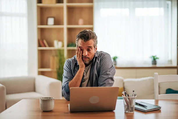 Tired businessman sitting at a dining table, working from home, feeling overwhelmed and bored, struggling with fatigue and stress while facing daily challenges of remote work