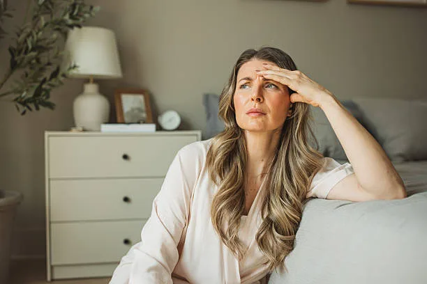 Mature menopause woman waking up. She is sitting in bedroom in pajamas and having headache.